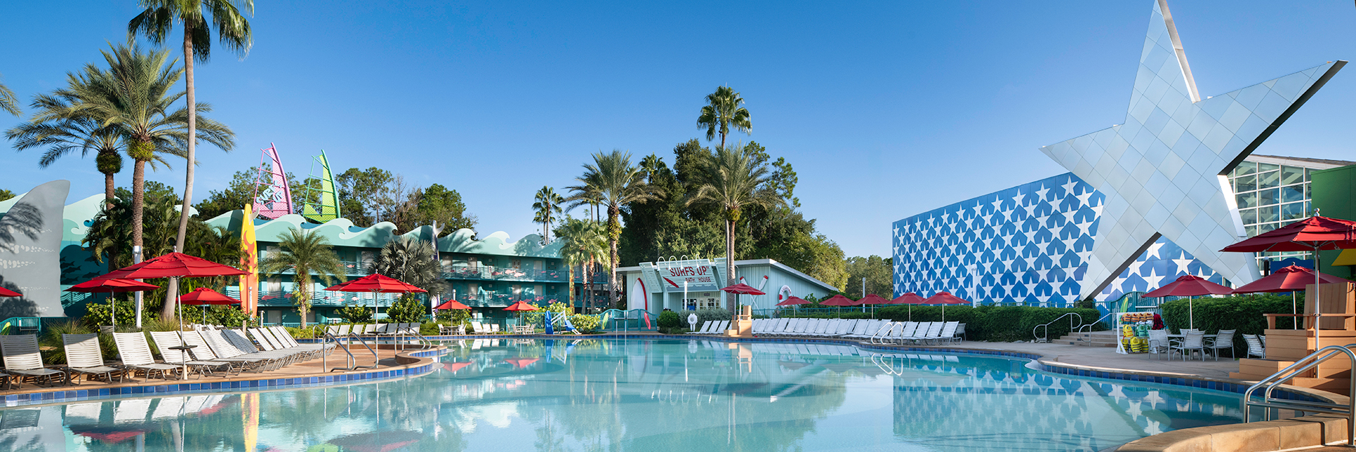 Wide view of an All Star Sports Resort pool with palm trees, lounge chairs, and colorful, themed buildings one with a large star