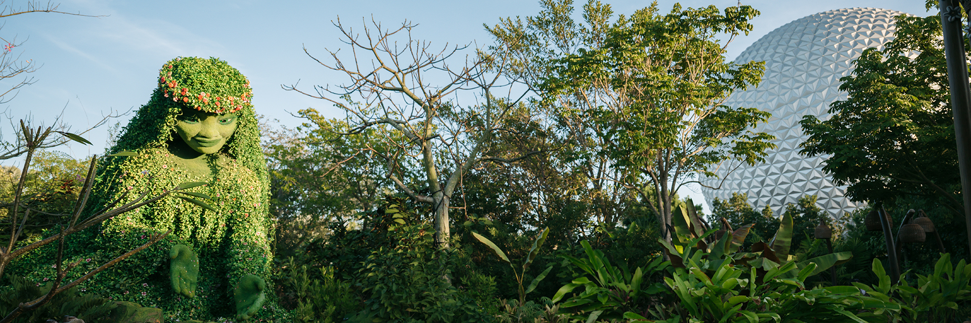 A lush garden scene featuring an oversized Te Fiti sculpture made of plants, with trees and a EPCOT's Spaceship Earth in the bac